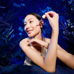 Woman applying Manuka Health Pro Vitality Face Serum skincare product with a dropper against a dark, nature-themed background.