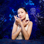 Woman holding Manuka Health Pro Vitality Face Cream skincare product with 'ECOCERT Cosmos Natural' logo against a blue floral background.
