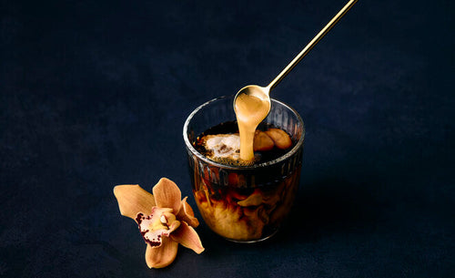 A dessert in a clear glass cup with a spoon pouring caramel-like sauce over it, set on a dark navy background with a peach orchid flower beside it.