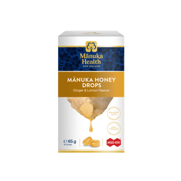 A box of Mânuka Health Mânuka Honey Drops (Ginger & Lemon Flavour) on a solid white background. The packaging is white with a dark blue band at the top, a gold banner, and an illustration of honey dripping from a piece of ginger root.