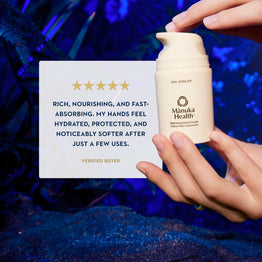 A person's hands hold a bottle of Manuka Health Pro Vitality Moisturizing Hand Cream next to a white card with a verified buyer review, set against a dark blue, textured background.